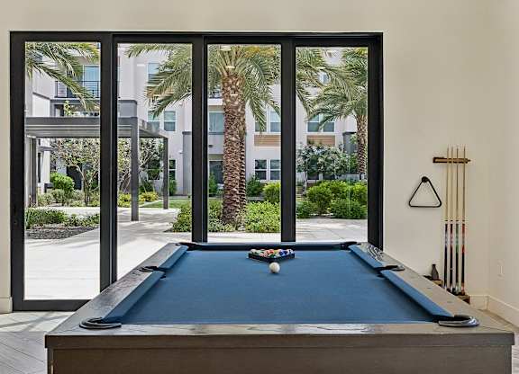 A pool table with a blue cloth and white balls is in the foreground of a room with a view of a palm tree and a building outside the window.