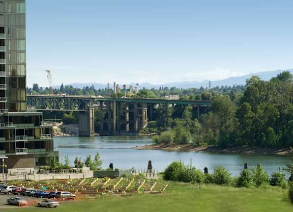 A river flows under a bridge with a building on the left and a parking lot in the foreground.