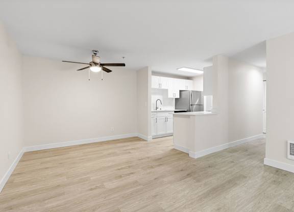 A room with a ceiling fan and light fixture, and a kitchen area in the background.