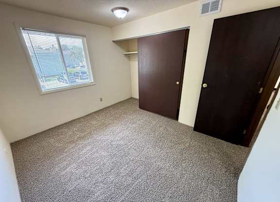 A room with a carpeted floor, a window with blinds, and two brown doors. at Winding Hills Apartments, Iowa