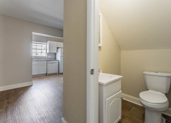 A white toilet sits in a bathroom next to a kitchen.
