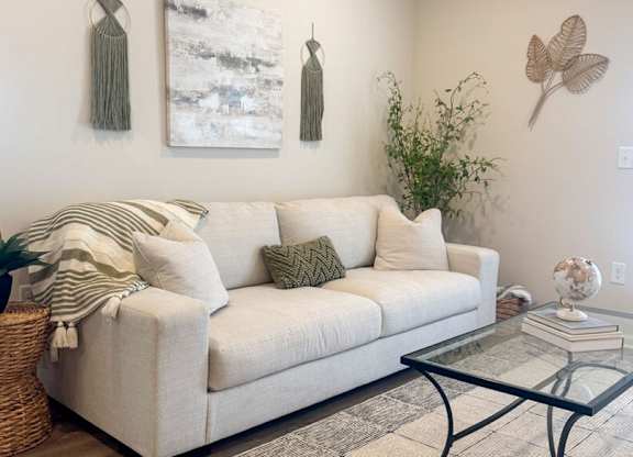 A living room with a white couch, a glass coffee table, and two wall hangings.