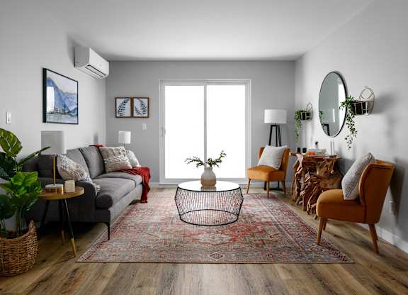 A living room with a grey couch, a red throw, a round table with a plant on it, a chair, a rug, and a mirror on the wall.