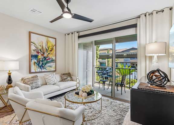 A living room with a white couch, a painting, a ceiling fan, and a glass door leading to a balcony.