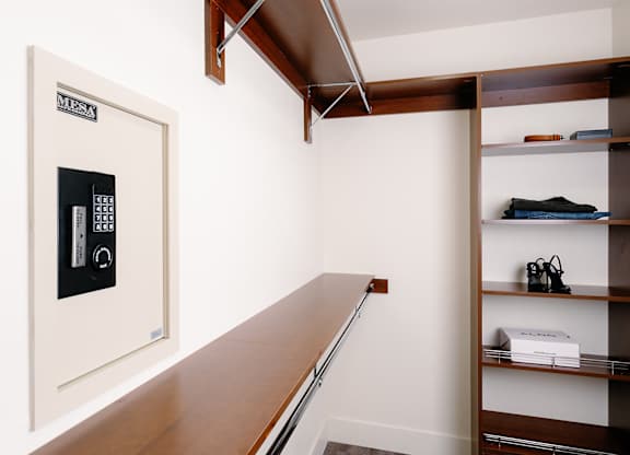 A white safe with a digital keypad is on the left side of a room with a wooden shelf on the right.