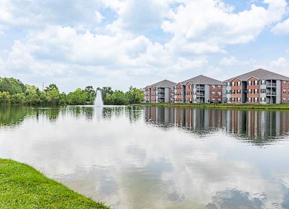 A serene lake with a fountain in the middle and apartment buildings in the background.
