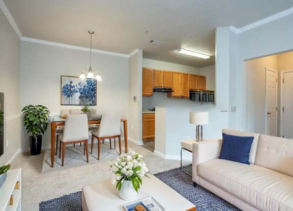 A living room with a white couch, a dining table with chairs, and a kitchen area in the background.