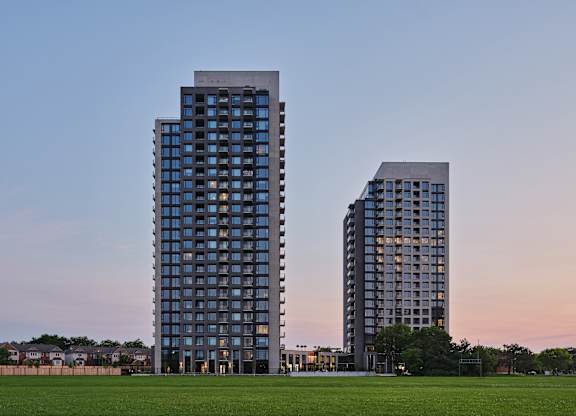 Two modern high-rise buildings stand side by side with a grassy field in the foreground.