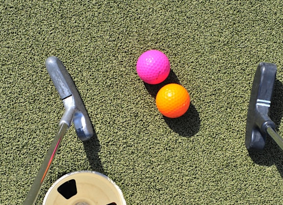 A golf ball, a small orange ball, and a pink ball are on a green surface with a golf club and a golf tee.