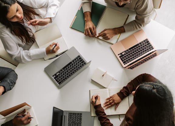 A group of people are sitting around a table with laptops and notebooks, appearing to be in a meeting or collaborative work session.