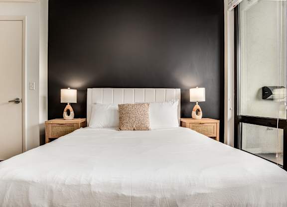 A bed with white bedding and two pillows sits in a room with a white door and a black wall.