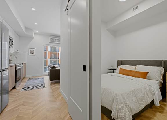 A bedroom with a bed, nightstand, and a kitchen area.