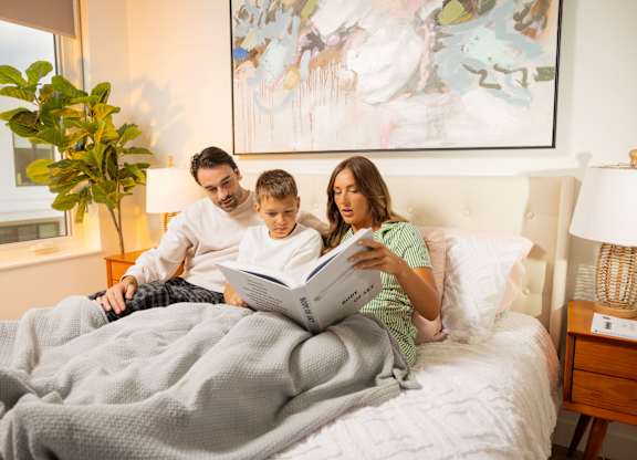 A family of three is reading a book together on a bed.
