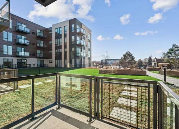 A balcony with a metal railing overlooks a grassy area and buildings.