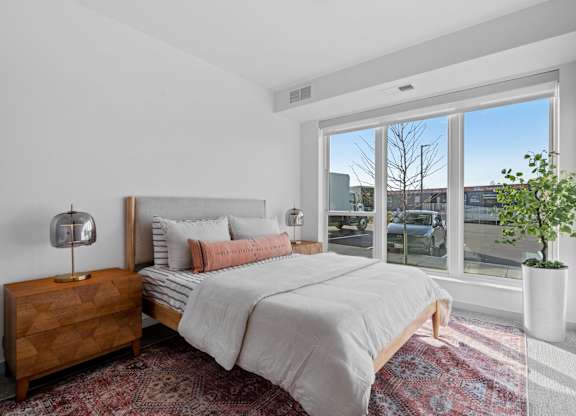 A bedroom with a large bed, a wooden nightstand, and a window overlooking a cityscape.