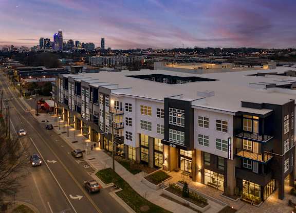 A modern apartment complex is lit up at dusk with cars driving on the road in front.