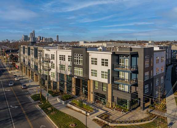 A modern apartment complex is situated on a street corner with a city skyline in the background.