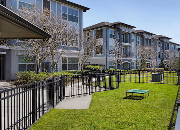 A black fence surrounds a grassy area with a bench in front of apartment buildings.