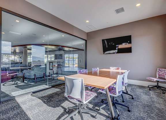 A conference room with a long table and chairs, a television on the wall, and a view of the outside through a glass wall.