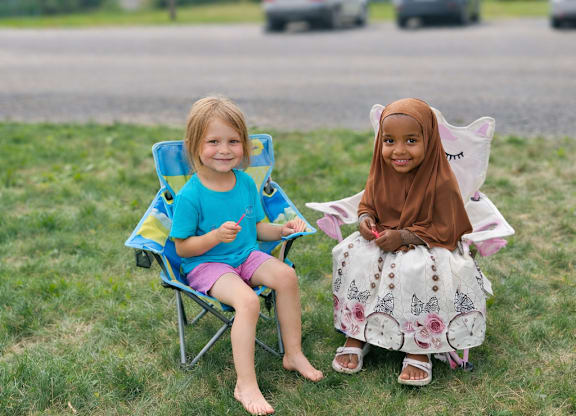Two children sitting on chairs on the grass.