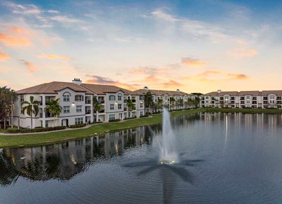 A fountain in the foreground of a serene lake in front of apartment buildings.