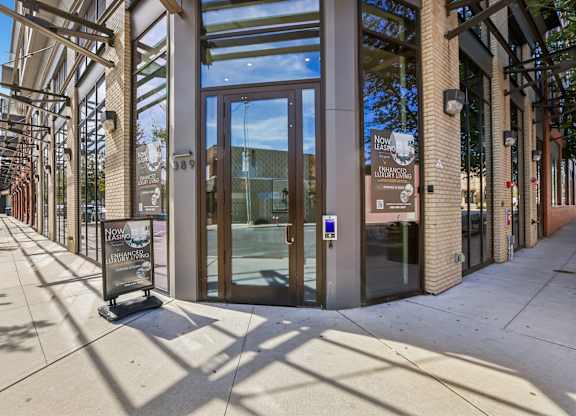 The image shows a modern building entrance with a glass door and a sign that reads "Now Open".