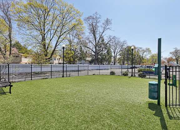 A park with a green grass field, a bench, a trash can, and a fence.