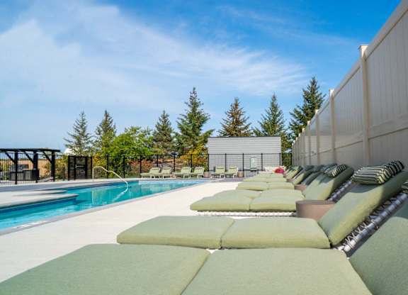 A row of green cushioned lounge chairs are arranged on a patio next to a pool.