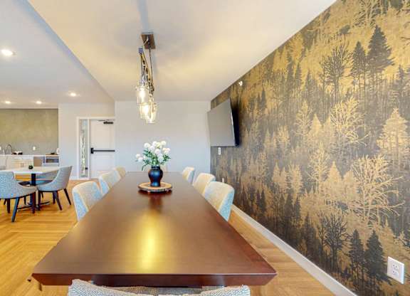 A long wooden table is surrounded by chairs in a room with a forest wallpaper.