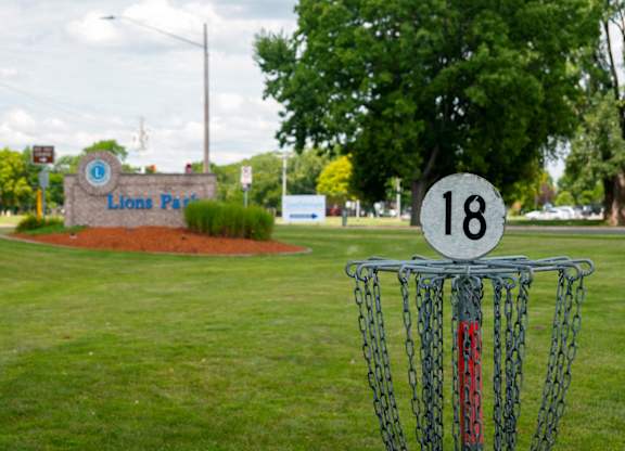A disc golf basket with the number 18 on it is in the foreground of a grassy area with a sign that says Lions Park in the background.