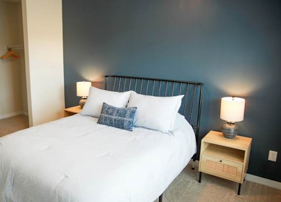 A bed with a white comforter and a blue pillow is in a room with a lamp on a nightstand.
