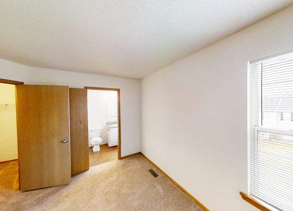 A room with a carpeted floor, a window with blinds, and a door leading to a bathroom.