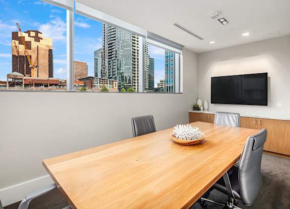 A conference room with a wooden table and chairs, a television, and a view of the city skyline through the windows.