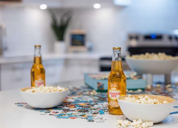 Two bottles of beer are on a table with bowls of popcorn and puzzle pieces scattered around.