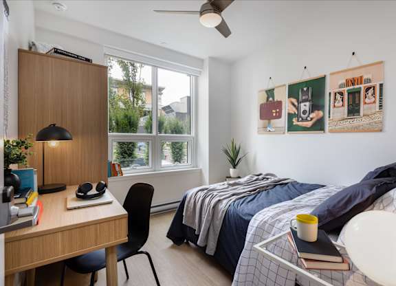 A bedroom with a bed, desk, chair, and a window with a view of trees and buildings outside.