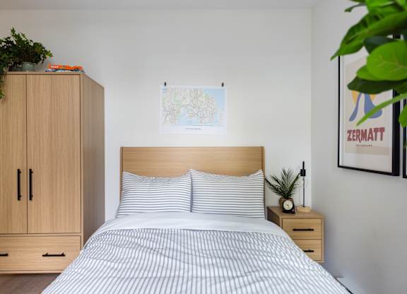 A bed with a wooden headboard and striped bedspread sits in a room with a wooden cabinet and a plant.