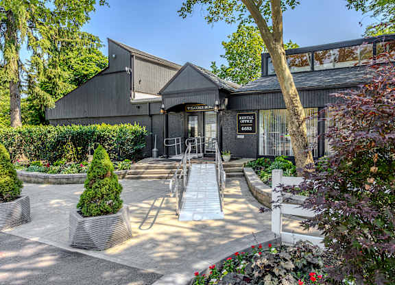 A building with a dark grey exterior and a black roof is surrounded by a well-manicured garden with a variety of plants and trees. at Commons At Waters Edge, Columbus Ohio