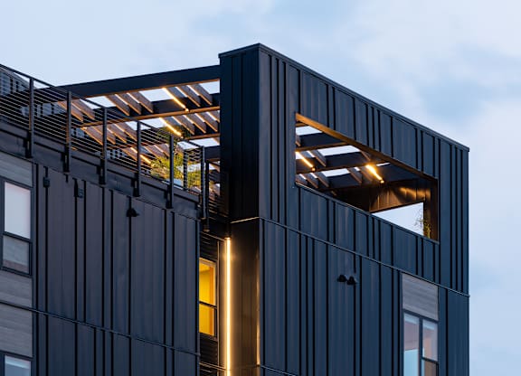 A modern building with a dark facade and a large window with a light shining through.
