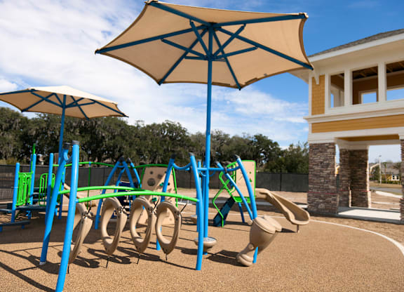 A playground with a blue and yellow swing set and a tan umbrella.