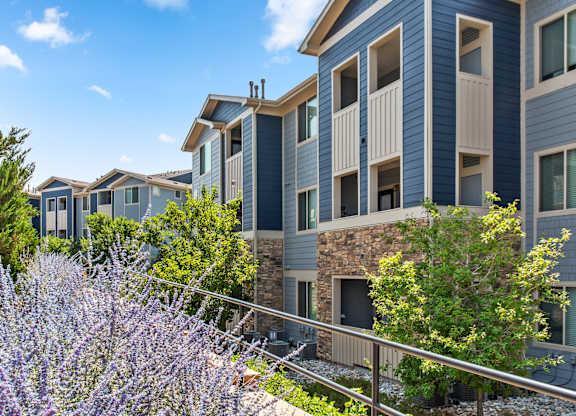 A row of modern apartment buildings with a fence and purple flowers in the foreground.