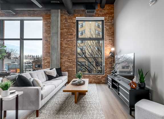 A living room with a grey couch, a wooden coffee table, and a large window with a view of a tree.at Raymond Block, Edmonton Alberta T6E 3R7