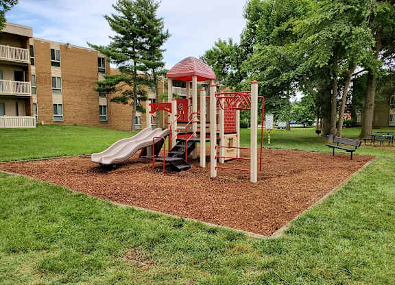 a playground with slides and monkey bars in a grassy area with a brick building in the at The Glendale Residence, Maryland, 20706