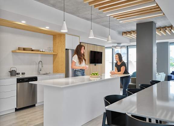 Two women are standing in a modern kitchen with a white countertop and wooden accents.