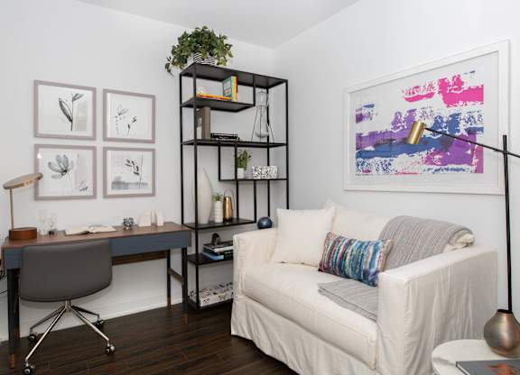 A living room with a white couch, a desk with a chair, and a bookshelf with a plant on top.