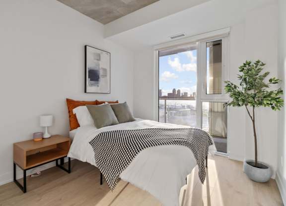 A bedroom with a bed, a nightstand, and a window overlooking a cityscape.