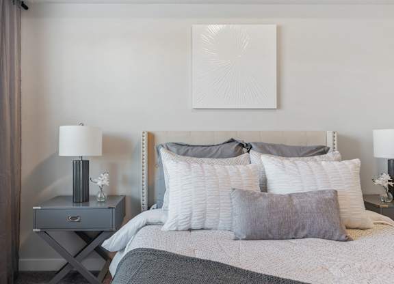 a bedroom with a bed and pillows and a nightstand with a lamp and at The Flats at Riverwoods, Provo, UT, 84604