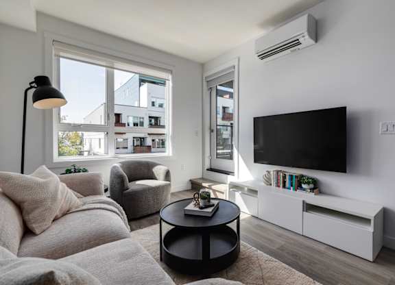 A living room with a large window, a grey sofa, a black coffee table, a white TV stand, and a floor lamp.