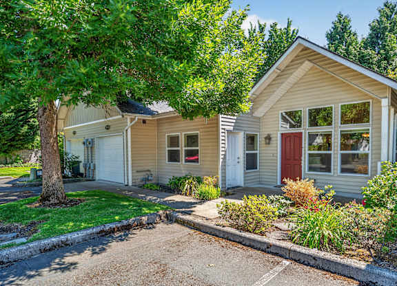 A house with a red door and a garage door is surrounded by greenery.