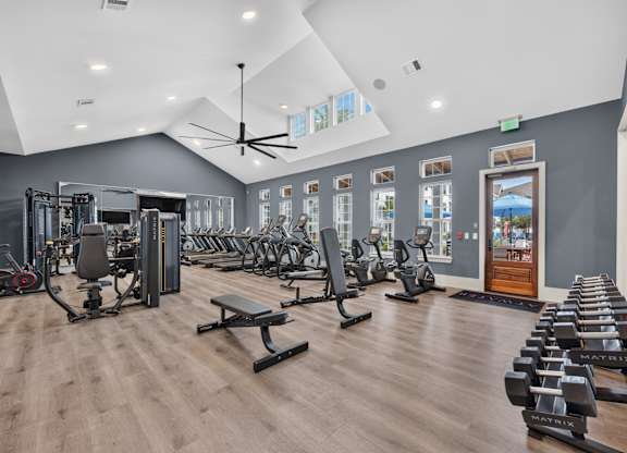 A gym with a variety of equipment including treadmills, weights, and a bench press. Large well lit room with a fan and tvs
