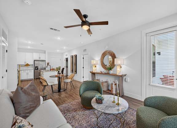 A living room with a grey couch, a green armchair, and a coffee table, fan with a lot of natural light, the kitchen and patio are also viewable in the image